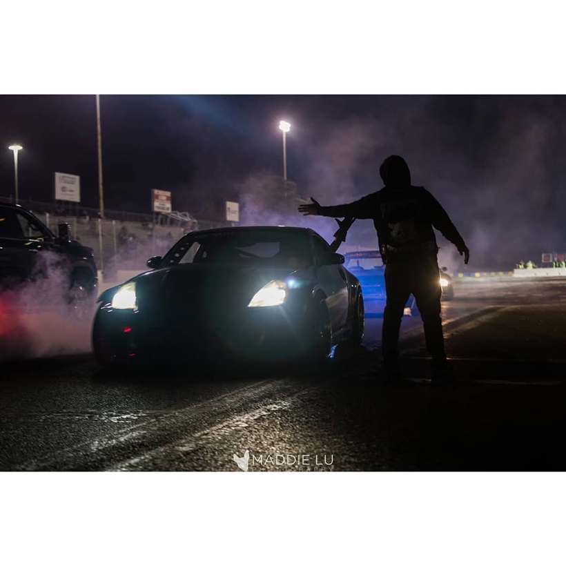 Night session — headlights cutting through tyre smoke as a grid marshal waves cars out.