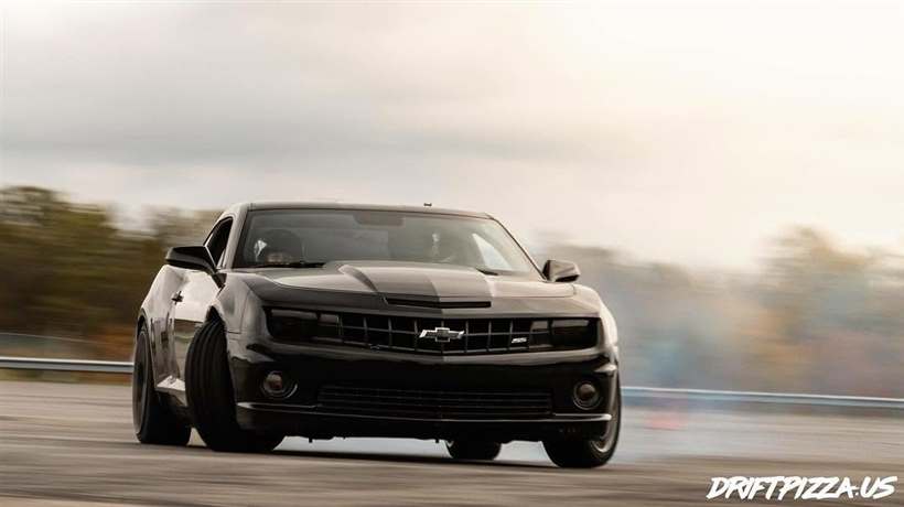 Lucas Prisuta's black Camaro SS sliding on a cold overcast day, autumn trees blurred behind it.