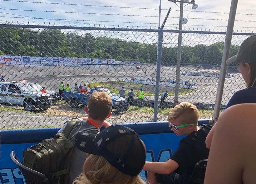 Family watching a session from the grandstand through the catch fence on a bright summer day.