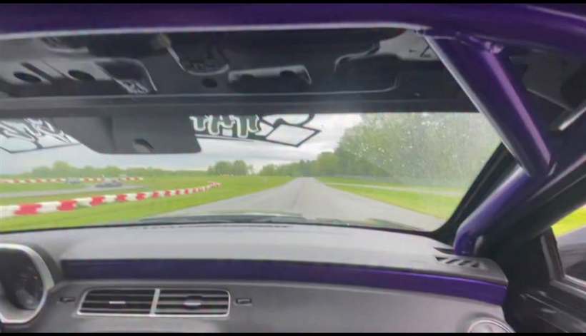 View from inside the car through the windscreen: purple roll cage, rumble strips and green grass ahead.