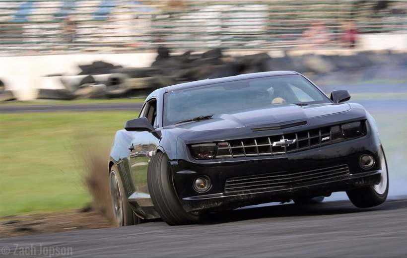 The Camaro dropping a wheel off the asphalt, throwing dirt, front wheels turned hard into the slide.